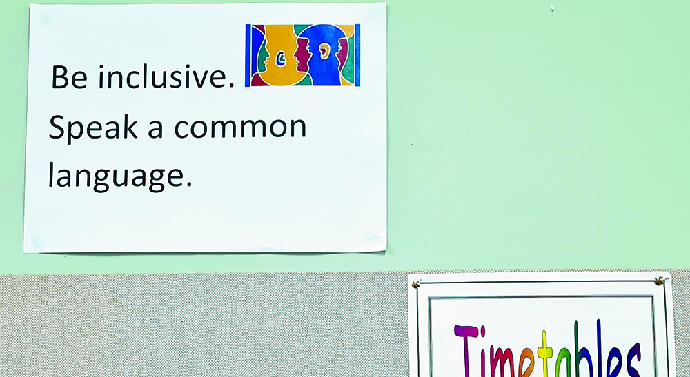 A sign on a wall with the text "Be inclusive. Speak a common language." alongside a colorful illustration of faces in profile, above another sign labeled "Timetables."