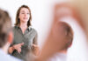 Speak with power: embodied confidence for pronunciation skills A woman in a green shirt is speaking in front of an audience, gesturing with her hands, and appears focused on delivering her presentation. Blurred figures of audience members are visible in the foreground, suggesting an interactive session.