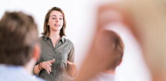 A woman in a green shirt is speaking in front of an audience, gesturing with her hands, and appears focused on delivering her presentation. Blurred figures of audience members are visible in the foreground, suggesting an interactive session.