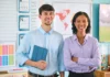 Two for the price of one! Two smiling teachers stand side by side in a classroom, with a young man in a light blue shirt holding a tablet and a woman in a lavender blouse crossing her arms. Behind them, educational posters and a world map are visible on the walls.