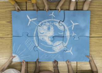 Rethinking sustainability: it starts with being self-sustainable Children assembling a large blue puzzle featuring a chalk drawing of Earth with symbols of renewable energy, including wind turbines and solar panels.