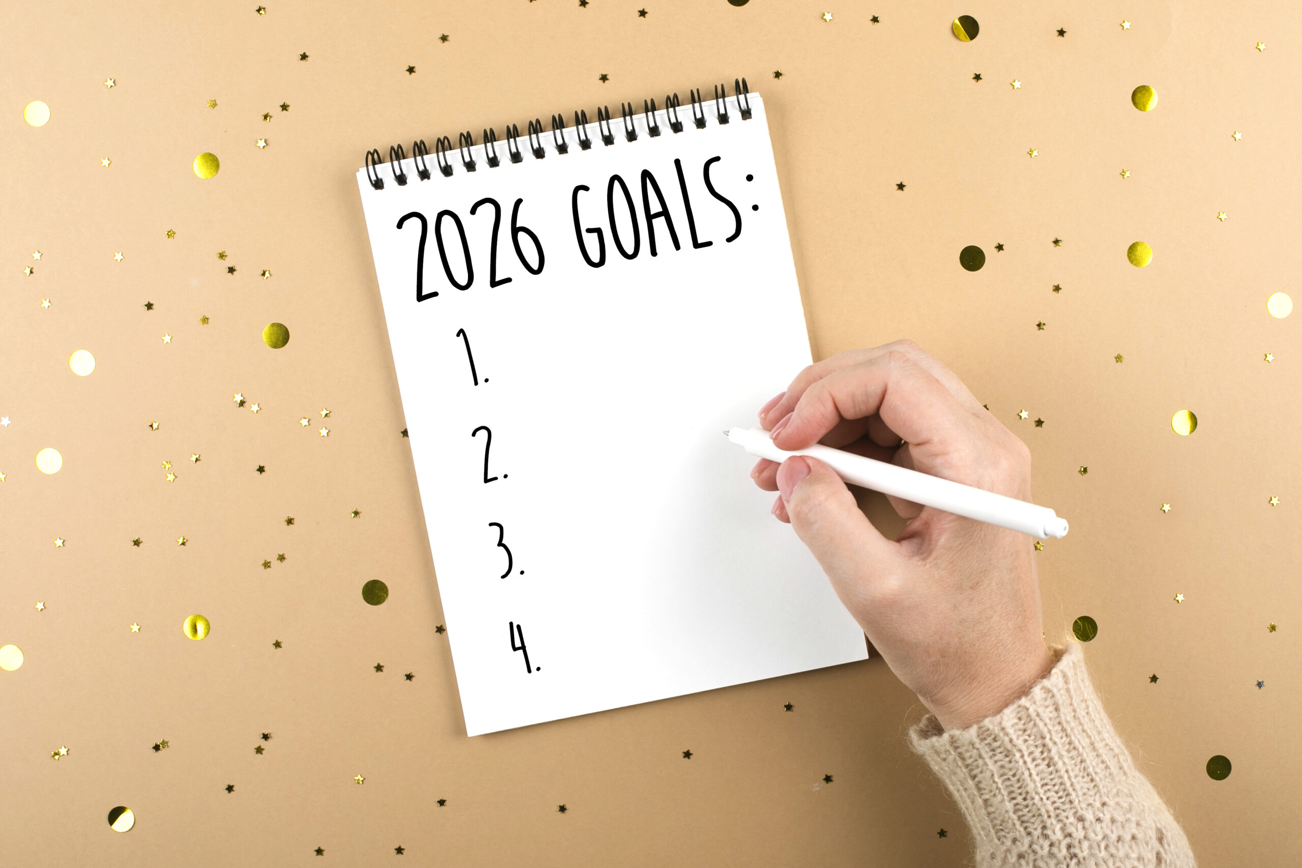 A person holding a pen poised to write on a spiral notebook, with the words '2026 Goals:' followed by a numbered list from 1 to 4 on a blank page, set against a beige background with scattered gold star and circle confetti.
