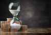 Be wise – Time is money An hourglass with sand flowing inside, surrounded by stacks of coins placed on a wooden surface, against a dark blurred background.