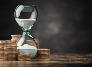 Be wise – Time is money An hourglass with sand flowing inside, surrounded by stacks of coins placed on a wooden surface, against a dark blurred background.