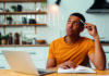 Which barriers to creativity do school leaders need to be aware of? A person in an orange shirt sitting at a desk with a laptop and an open book, holding a pen to their temple and looking up thoughtfully.