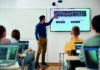 The complete integration of pronunciation with everything else A teacher in a blue shirt and beige trousers is pointing at a large screen displaying lists of simple and extreme adjectives. Students seated at desks with laptops are attentively watching the presentation.