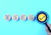 Conducting a criterion-referenced materials evaluation Four wooden discs with icons representing documents in a row, with a hand holding a magnifying glass over a yellow disc with a large checkmark against a light blue background.