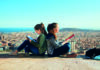 Travel readers Two girls sitting back-to-back, reading books on a rooftop with a panoramic view of a city skyline and the ocean in the background.