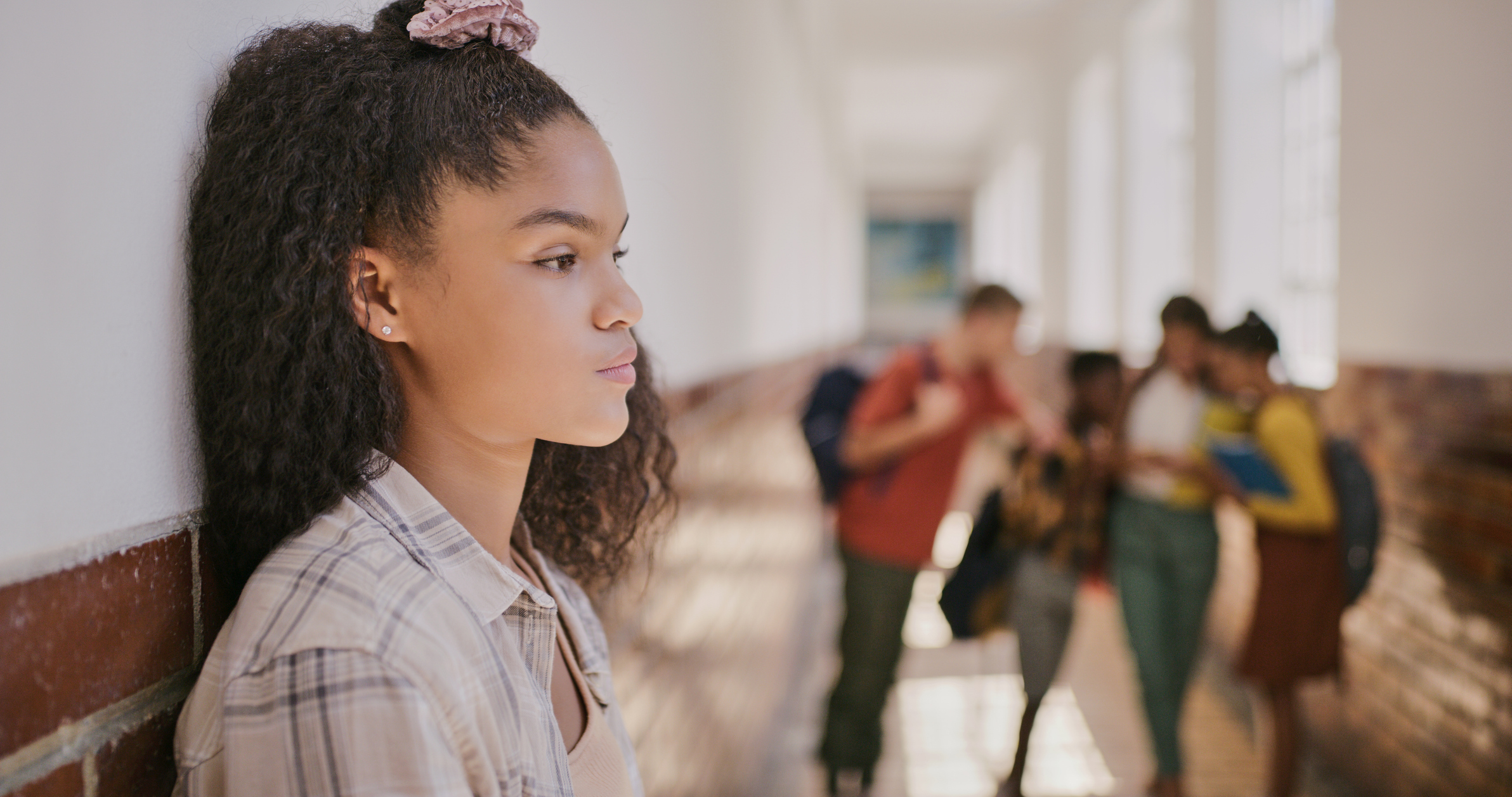 A young girl with curly hair and a scrunchie stands against a wall, looking thoughtful in a school corridor. In the background, a group of students are gathered, conversing with each other.