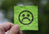 Shy or Unsafe? Why some learners struggle with speaking in the language classroom A hand holding a crumpled green sticky note with a sad face emoticon drawn on it, set against a blurred outdoor background.