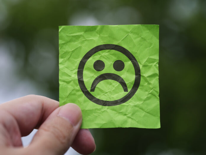 A hand holding a crumpled green sticky note with a sad face emoticon drawn on it, set against a blurred outdoor background.