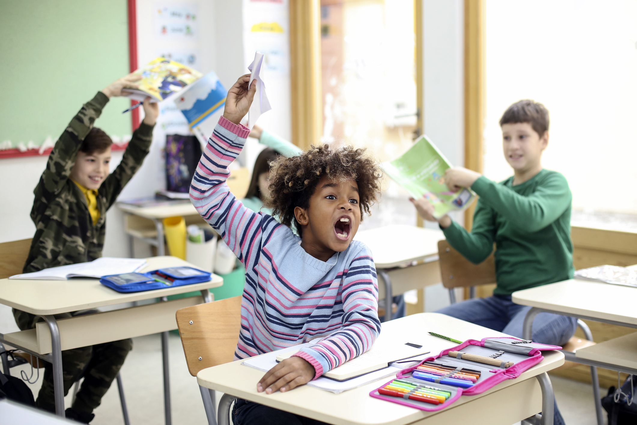 Children in a classroom enthusiastically participating, with one child holding up a paper airplane, surrounded by books and stationery on their desks.