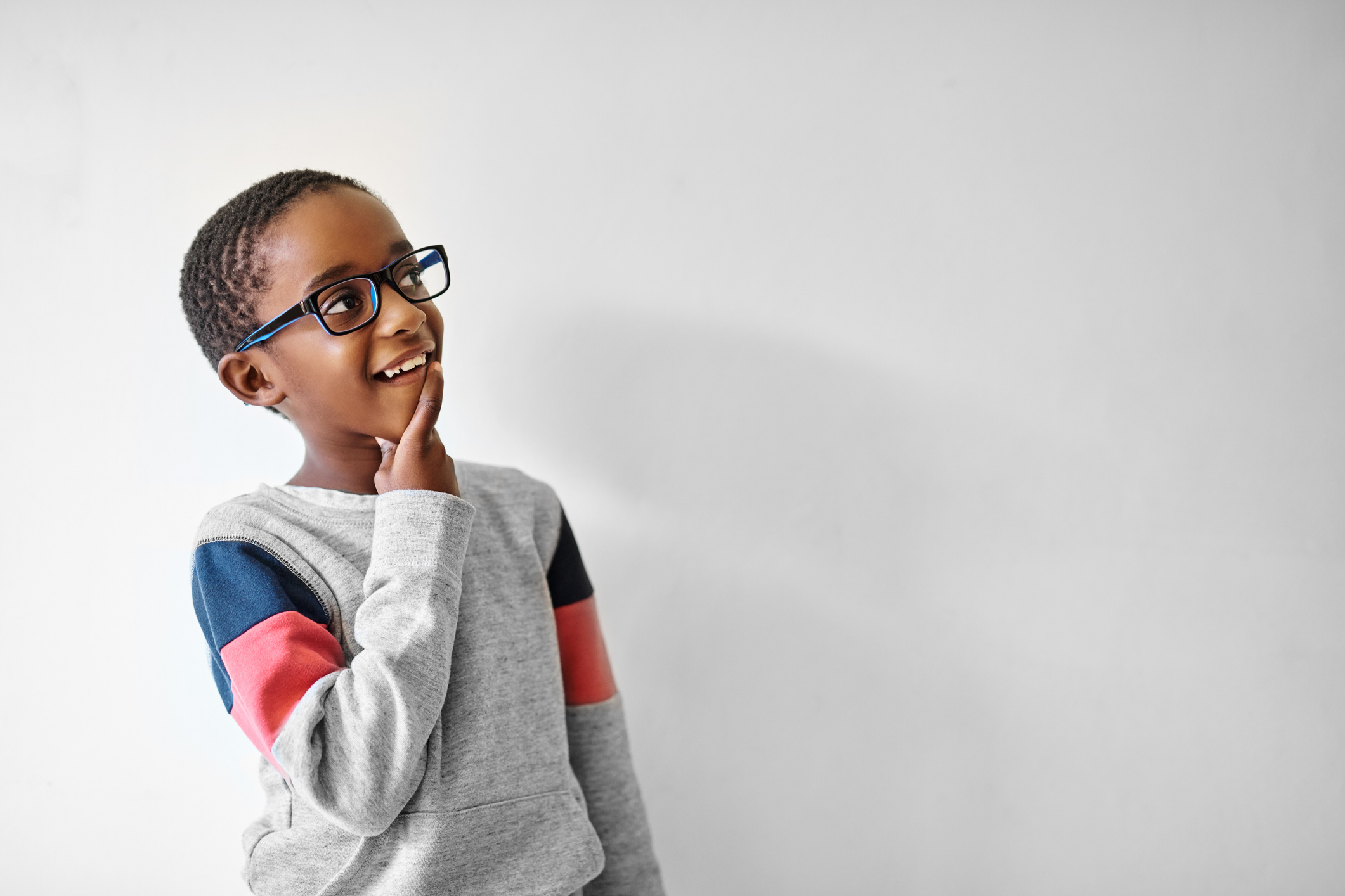 A young boy wearing glasses and a grey sweatshirt with colourful sleeves, standing against a white background, touching his chin and smiling thoughtfully.