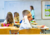 Managing the lot A young boy sitting at a desk in a classroom, resting his head on his hand and appearing lost in thought, with other children and a teacher in the background.