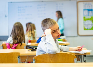 Managing the lot A young boy sitting at a desk in a classroom, resting his head on his hand and appearing lost in thought, with other children and a teacher in the background.