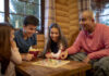 Things that amuse me A family of four playing a board game together in a cozy wooden cabin, with everyone smiling and one person holding a bowl of popcorn.