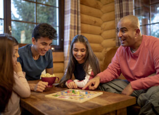 Things that amuse me A family of four playing a board game together in a cozy wooden cabin, with everyone smiling and one person holding a bowl of popcorn.