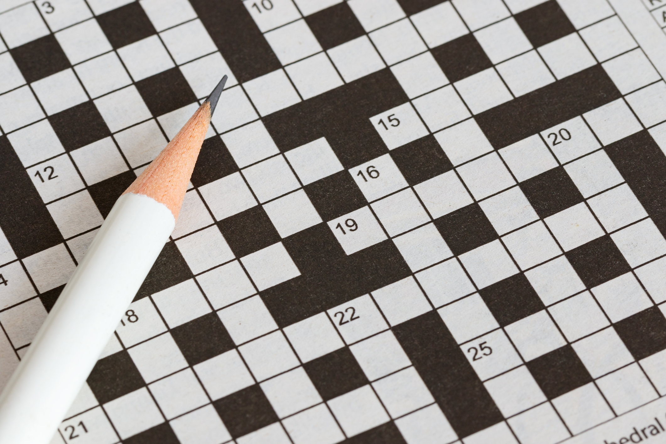A close-up image of a crossword puzzle with a sharpened pencil resting on it, showing blank squares and numbered clues.