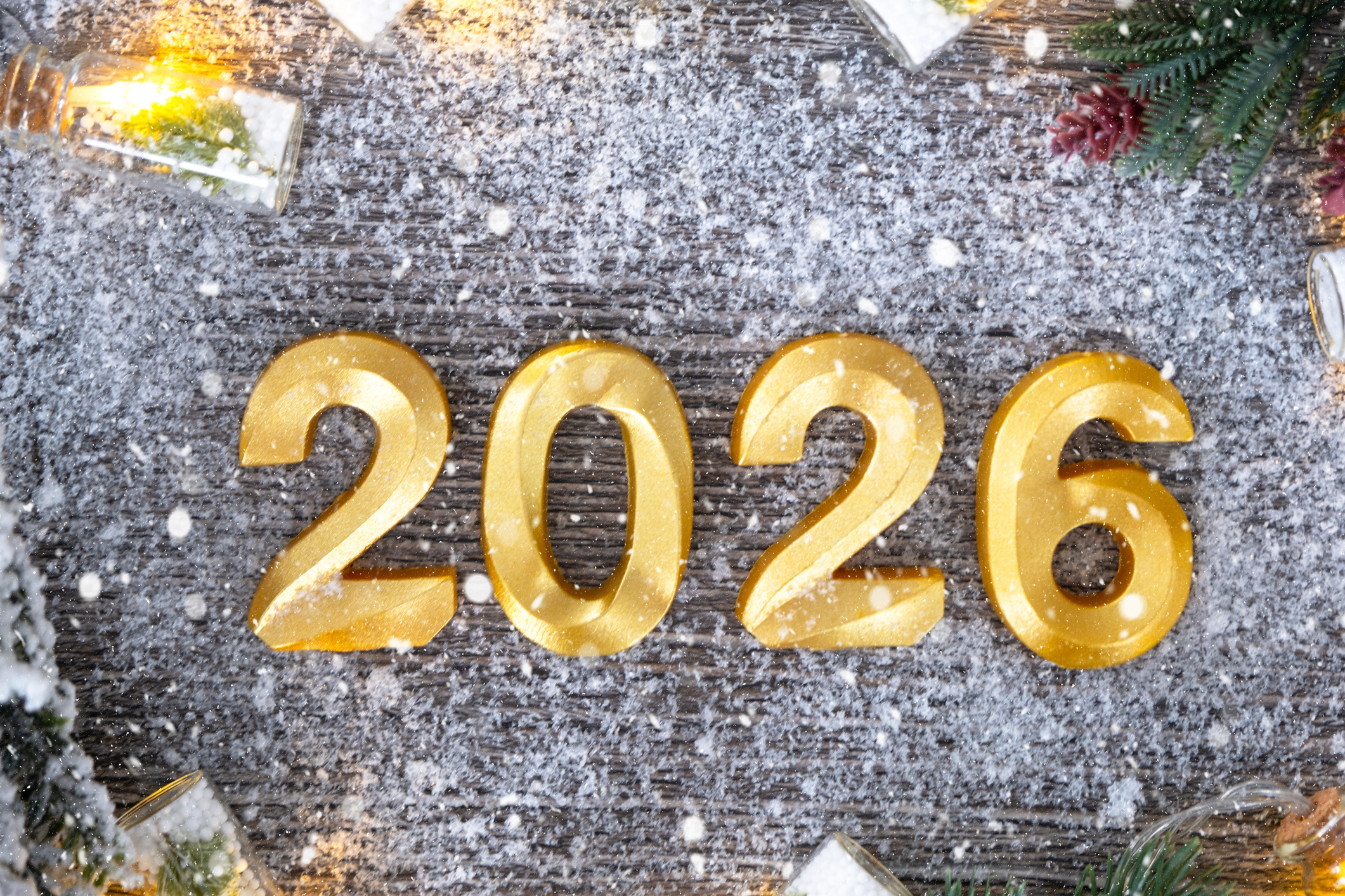 Gold numbers forming "2026" set on a wooden surface sprinkled with artificial snow, surrounded by festive elements like fir branches and small lights.