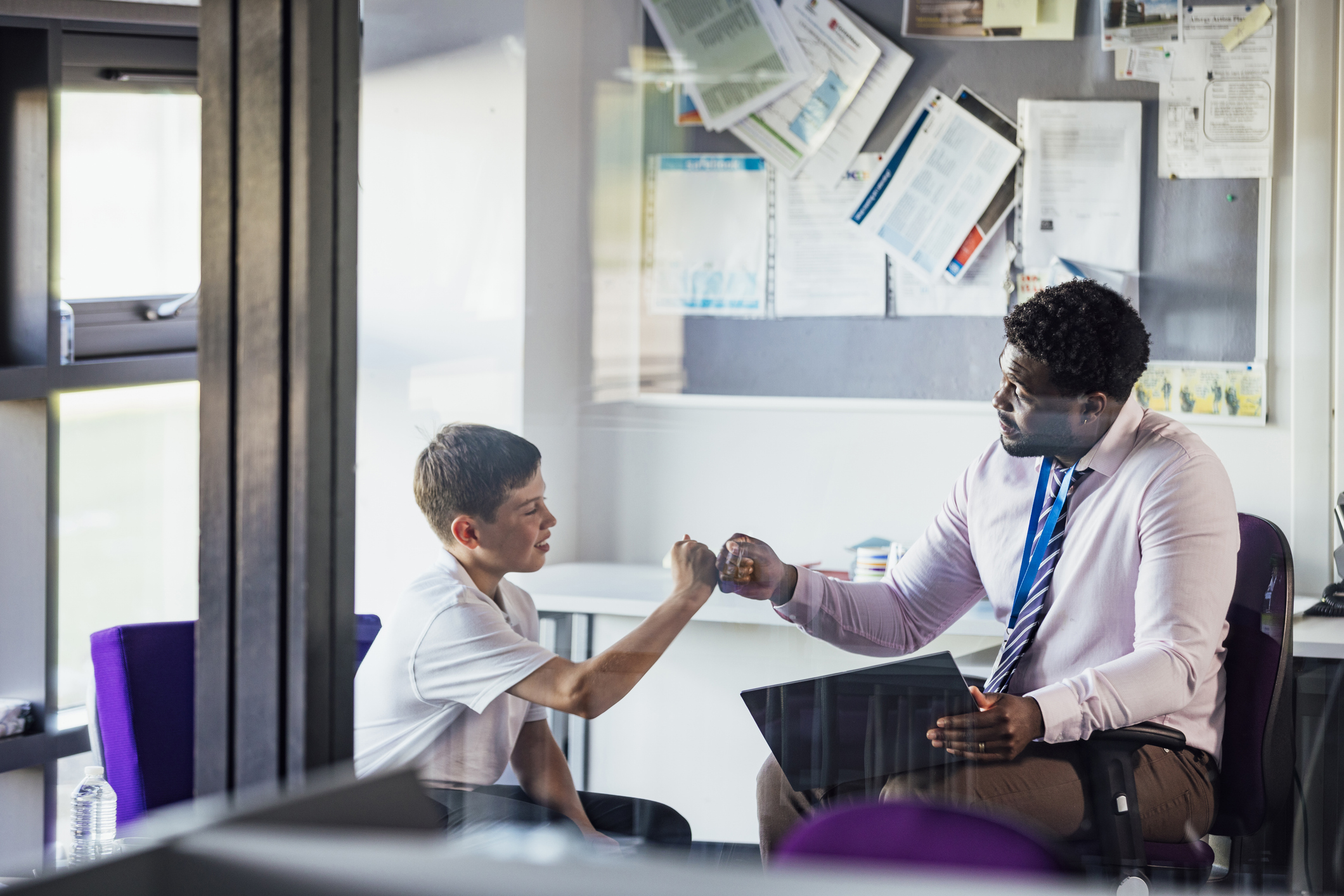A boy and a man seated in an office setting, giving each other a fist bump. The boy is wearing a white t-shirt, and the man is in a pink shirt with a blue tie, holding a folder. Papers and documents are pinned to a board in the background.