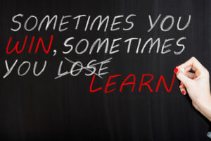A hand writing on a blackboard with chalk, showing the phrase 'SOMETIMES YOU WIN, SOMETIMES YOU LEARN,' with the word 'LOSE' crossed out and replaced by 'LEARN.'