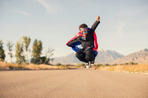 A young boy wearing a superhero costume, including a cape and mask, jumps on an empty road with mountains in the background under a clear sky.