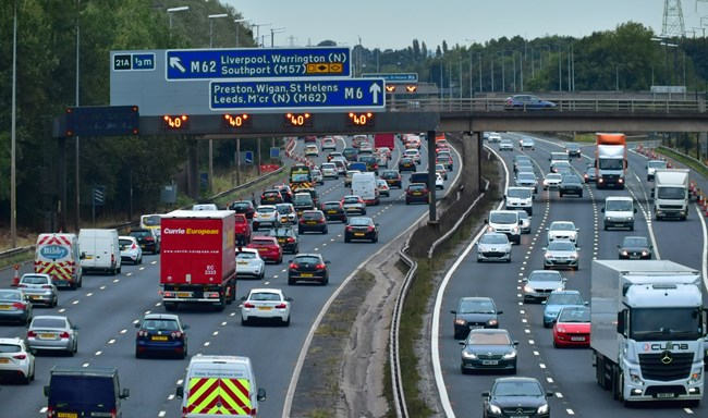 A busy motorway with multiple lanes of traffic, including cars and lorries, traveling under a large road sign. The sign directs traffic towards the M62 and M6, with destinations listed such as Liverpool, Warrington, Preston, and Leeds. There is a green landscape and trees in the background.