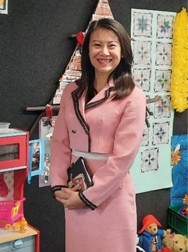 A woman in a pink suit with black trim smiling and standing in a room decorated with children's art and toys.