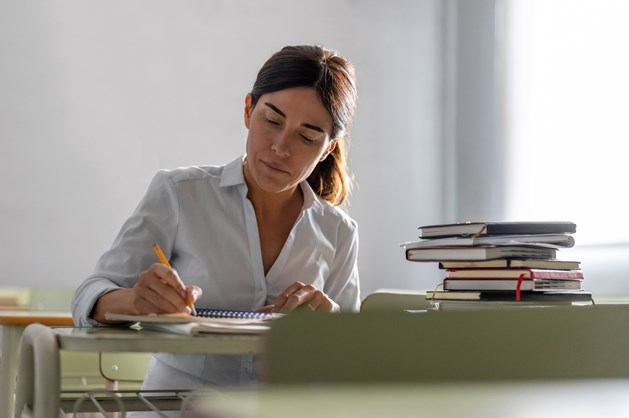A woman sitting at a desk, writing in a notebook with a pencil, next to a stack of books.