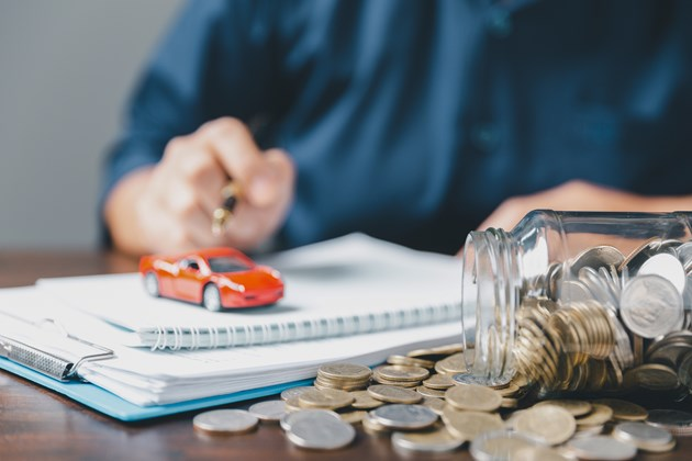 A person in a dark shirt sits at a desk with a pen in hand, next to a small red toy car on top of a notebook. A jar of scattered coins is in the foreground, suggesting themes of saving or financial planning for car expenses.