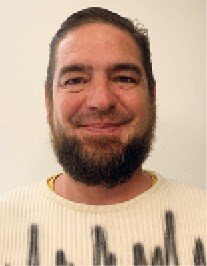 A person with a beard smiling at the camera, wearing a white sweater with black text.