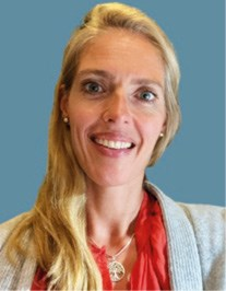 Smiling woman with long blonde hair, wearing a grey sweater over a red blouse, against a plain blue background.