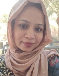 A woman wearing a light pink hijab and floral patterned outfit, smiling with a backdrop of palm trees.