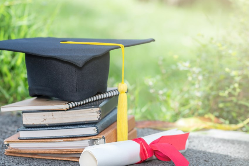 A graduation cap with a yellow tassel resting on a stack of books, accompanied by a rolled diploma tied with a red ribbon, set outdoors with grass in the background.