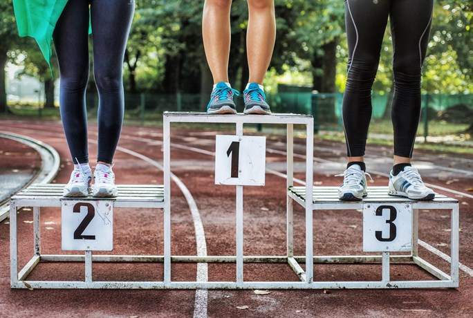 Three athletes standing on a winners' podium with numbers 1, 2, and 3 on a running track, showcasing their lower legs and feet.