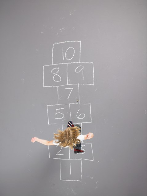 A child playing hopscotch on a chalk-drawn grid on a grey pavement, viewed from above.
