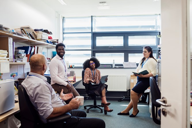 A group of four people having a meeting in a small office cluttered with books and papers. They are seated on chairs, engaged in conversation, with one person taking notes on a tablet or notebook. The office has a large window letting in natural light.