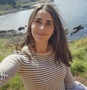 A woman with long brown hair wearing a striped shirt, taking a selfie with a scenic coastal background.