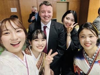 A group of five people smiling, with four women wearing traditional Japanese clothing and a man in a suit, posing indoors.