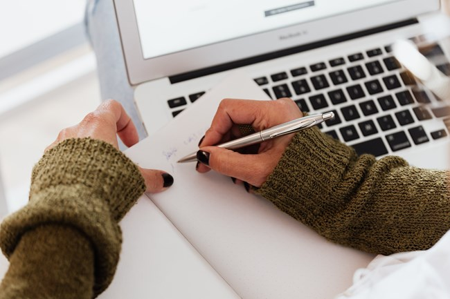 A person wearing a green knitted sweater is writing in a notebook with a silver pen near an open laptop.