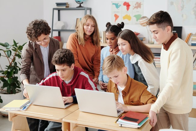A group of students gathered around two laptops in a classroom, with a teacher guiding them. The students are engaged and collaborating, surrounded by educational materials on shelves and maps on the walls.