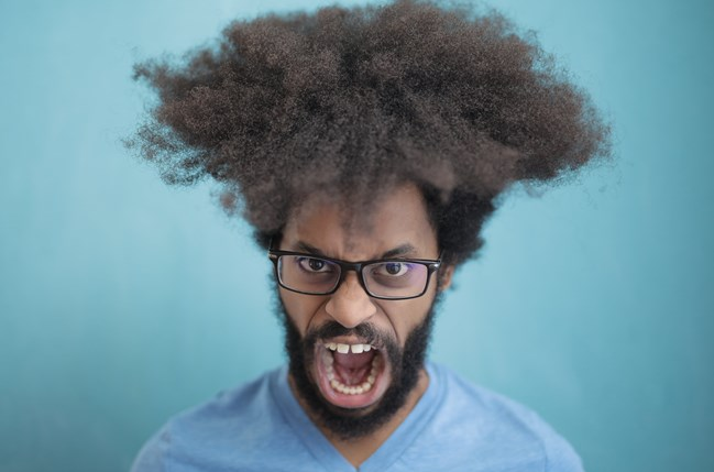 A person with a large, curly afro hairstyle and beard, wearing glasses and a blue shirt, appears to be shouting against a blue background.