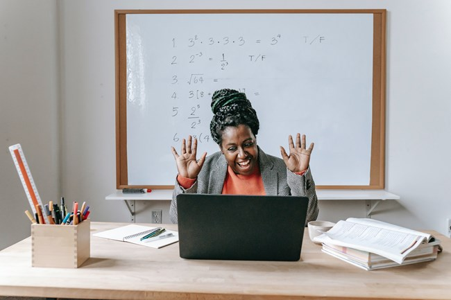 A person sitting at a desk with a laptop, raising their hands in excitement. Behind them is a whiteboard with mathematical equations. The desk has a variety of items, including books and a container of pencils.