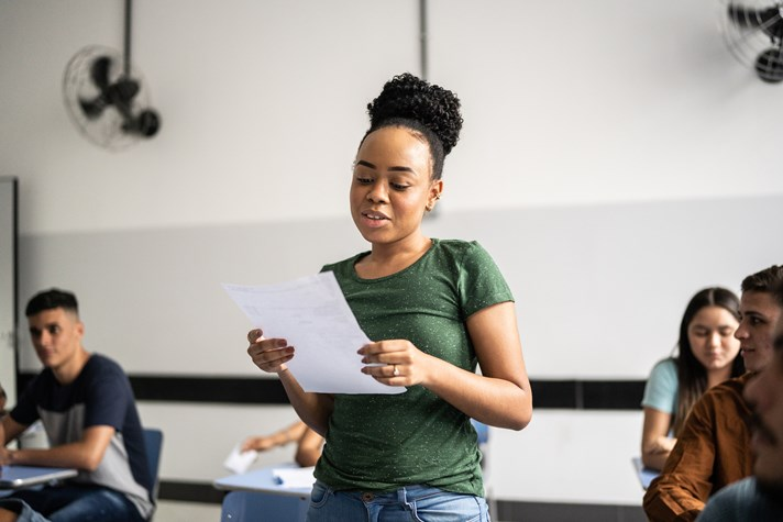 A woman stands in a classroom reading from a sheet of paper, while several other students sit in the background, engaged in various activities.