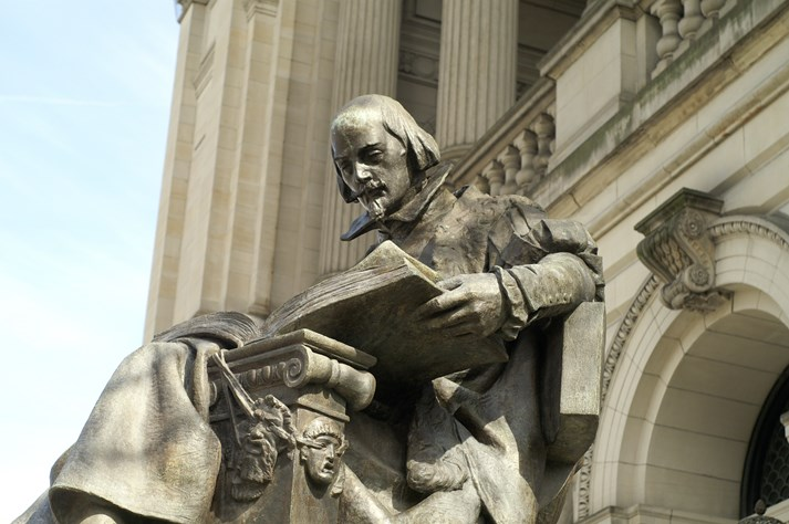 A statue of a historical figure reading a large book, set against the background of a grand building with pillars and ornate architecture.