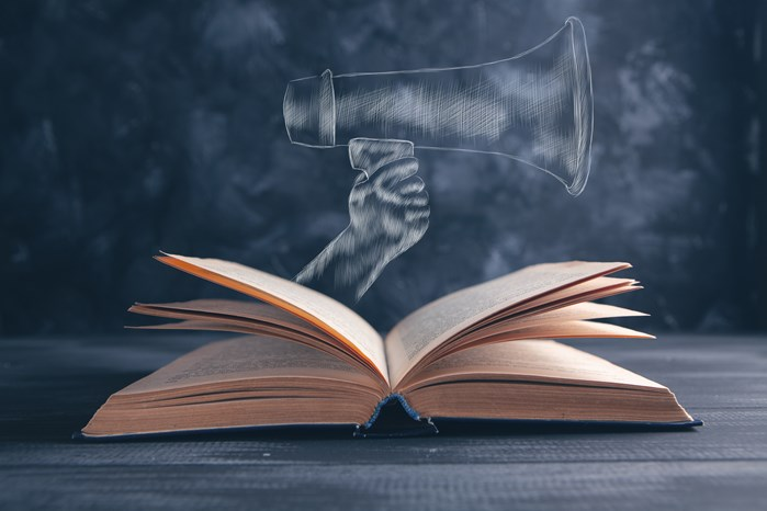 An open book with a chalk drawing of a hand holding a megaphone above it, symbolizing communication and knowledge sharing.