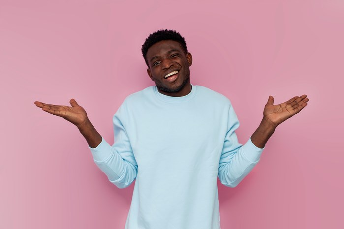 A person in a light blue sweater shrugging with both hands raised, standing against a pink background.