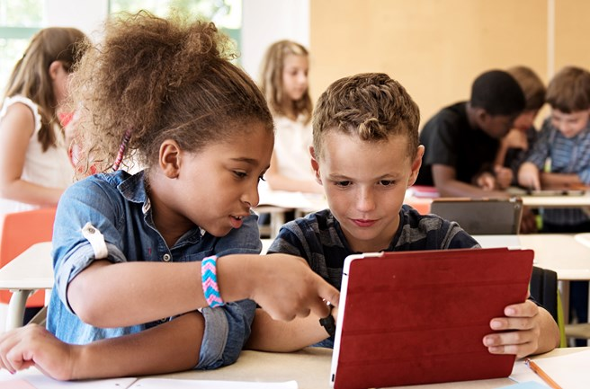 Two children sitting at a classroom desk, intently looking at a tablet screen; a girl points at the screen while a boy holds the device.