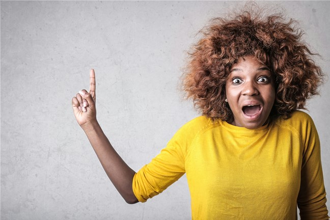 Person with curly hair wearing a yellow shirt, raising one finger while looking surprised.