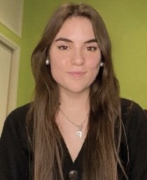 A person with long brown hair wearing a black top and a necklace, standing against a green wall.
